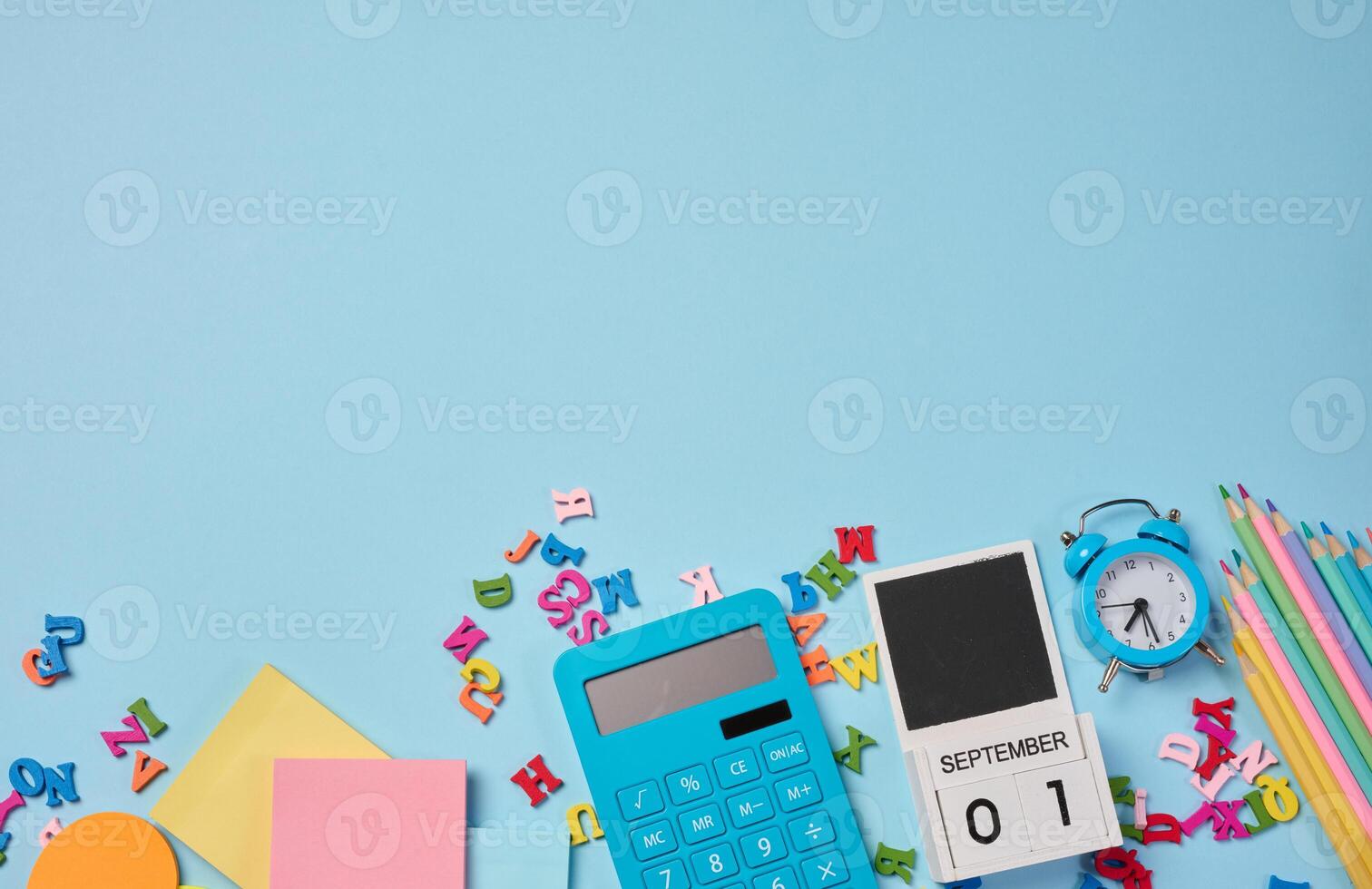 Stationery laid out on a blue background. Multi-colored pencils, calculator, alarm clock, sticky notes and wooden letters, preparing for study or work photo
