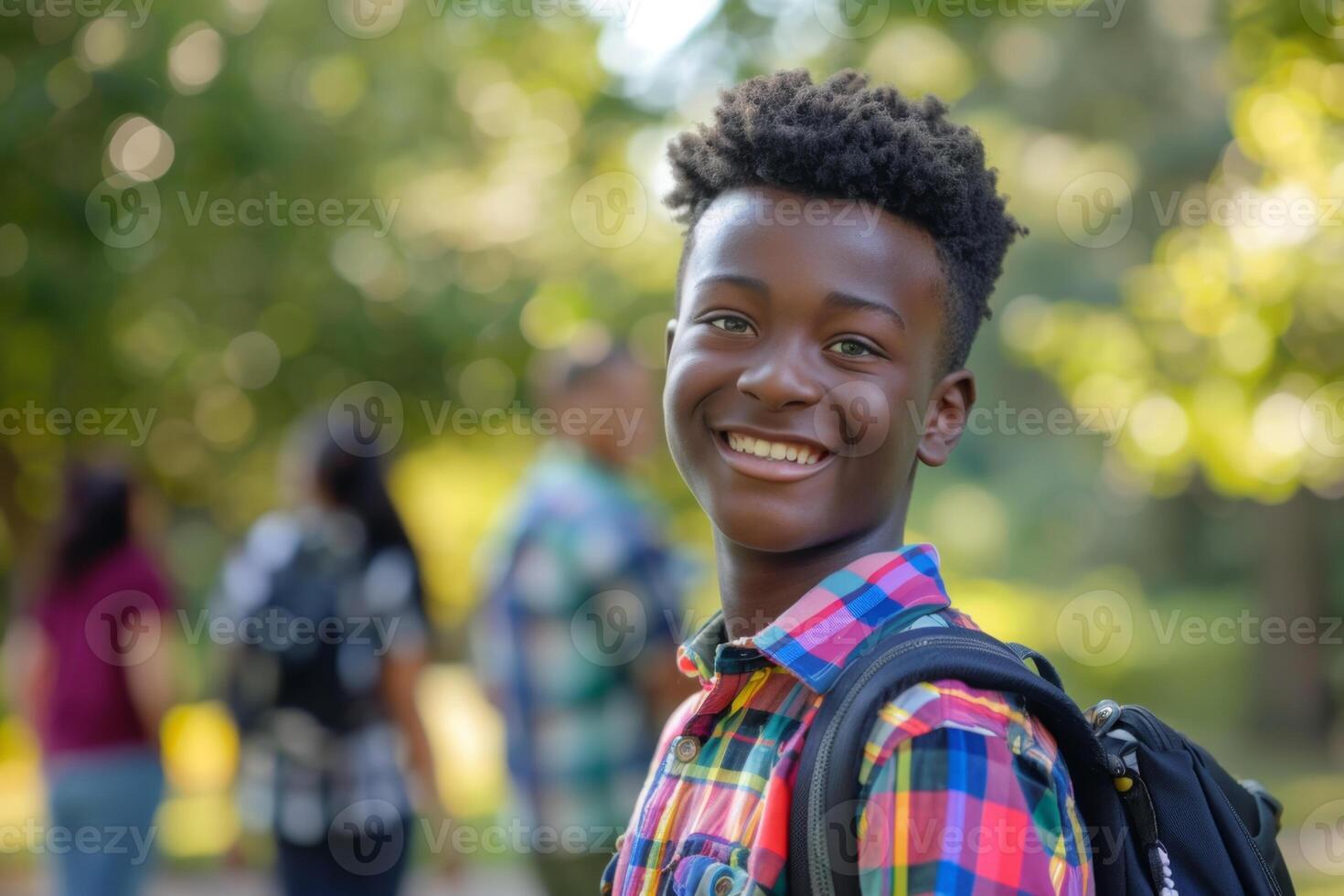 Smiling young black boy with short curly hair colorful plaid shirt carrying a backpack standing school outdoors blurred background back to school photo