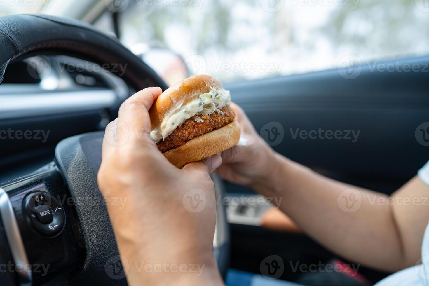 Asian lady holding hamburger to eat in car, dangerous and risk an accident. photo