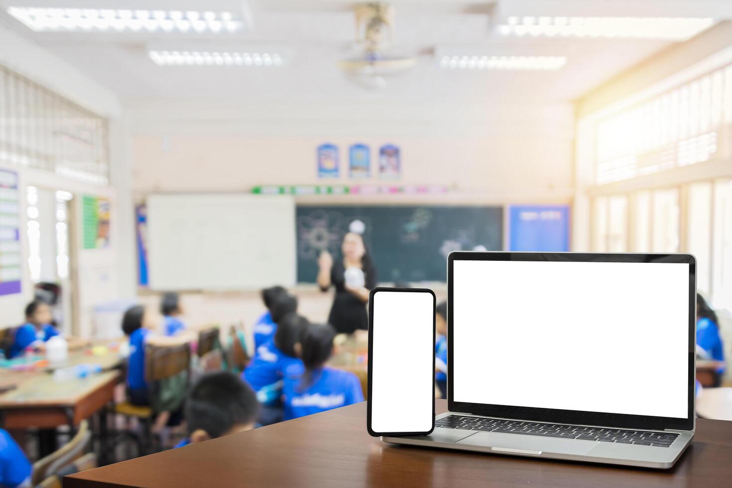 Front view, blank screen of black digital laptop and smartphone with tablet on the wood table. Children students in classroom background. photo