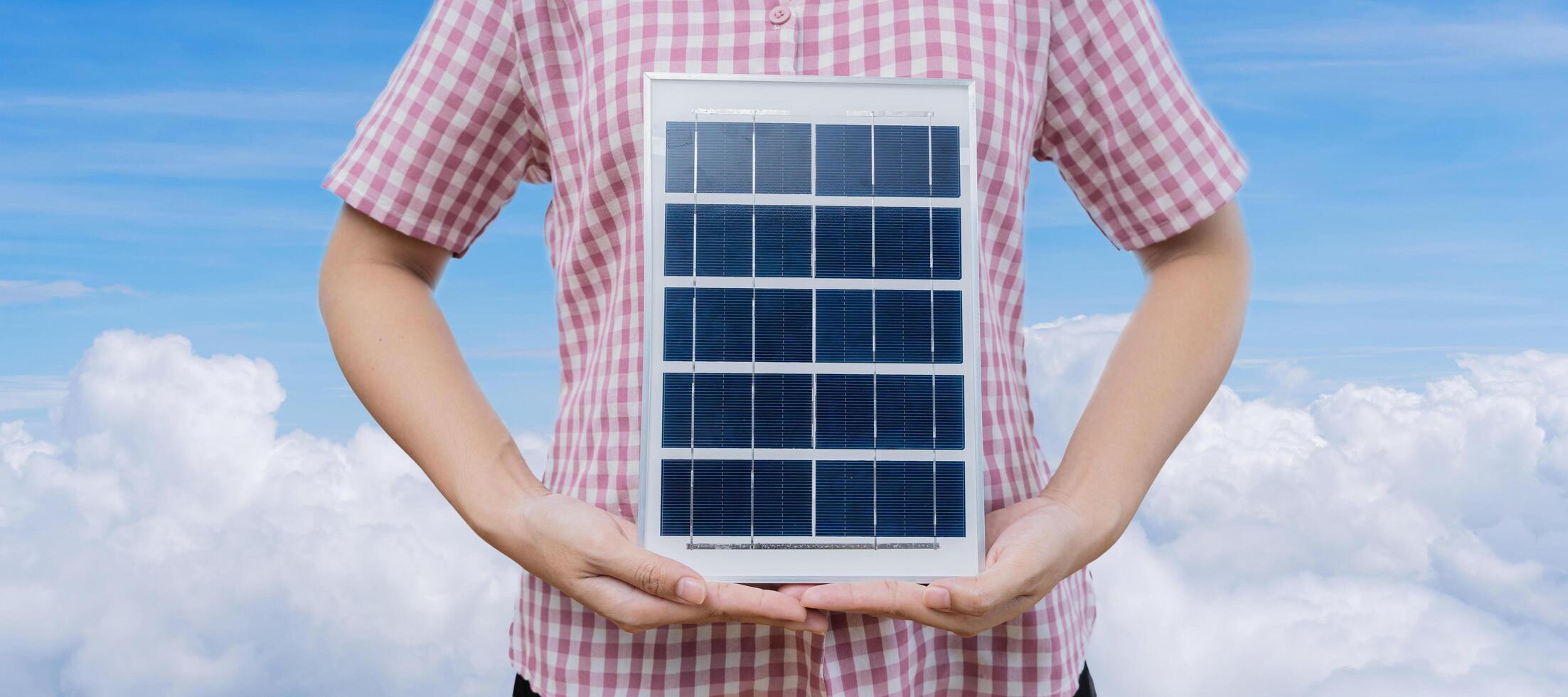 Successful woman holding solar panels and wind turbine with folded arms, isolated on blue sky background. Clean system technology and safety photo