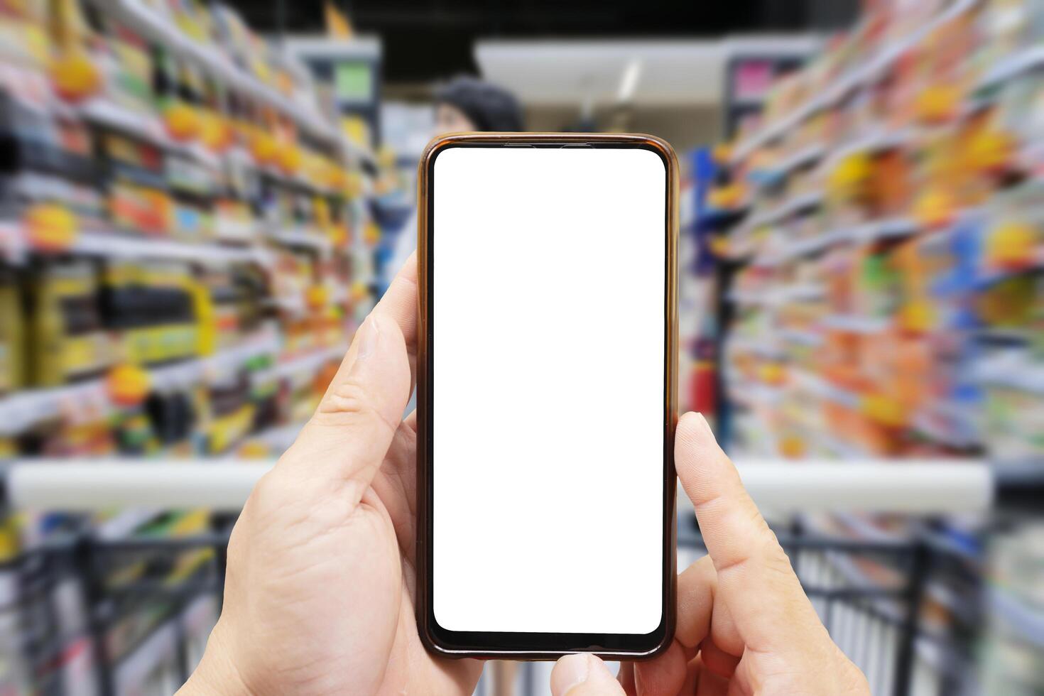 A person's hand holds a mobile phone, with a shopping basket on the screen. In the background, a pretty woman selects digital products, in a blur. Mock up. photo