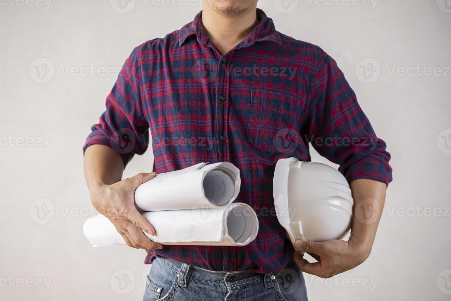 A young male engineer holding a blueprint with a white helmet while standing on a gray background. photo
