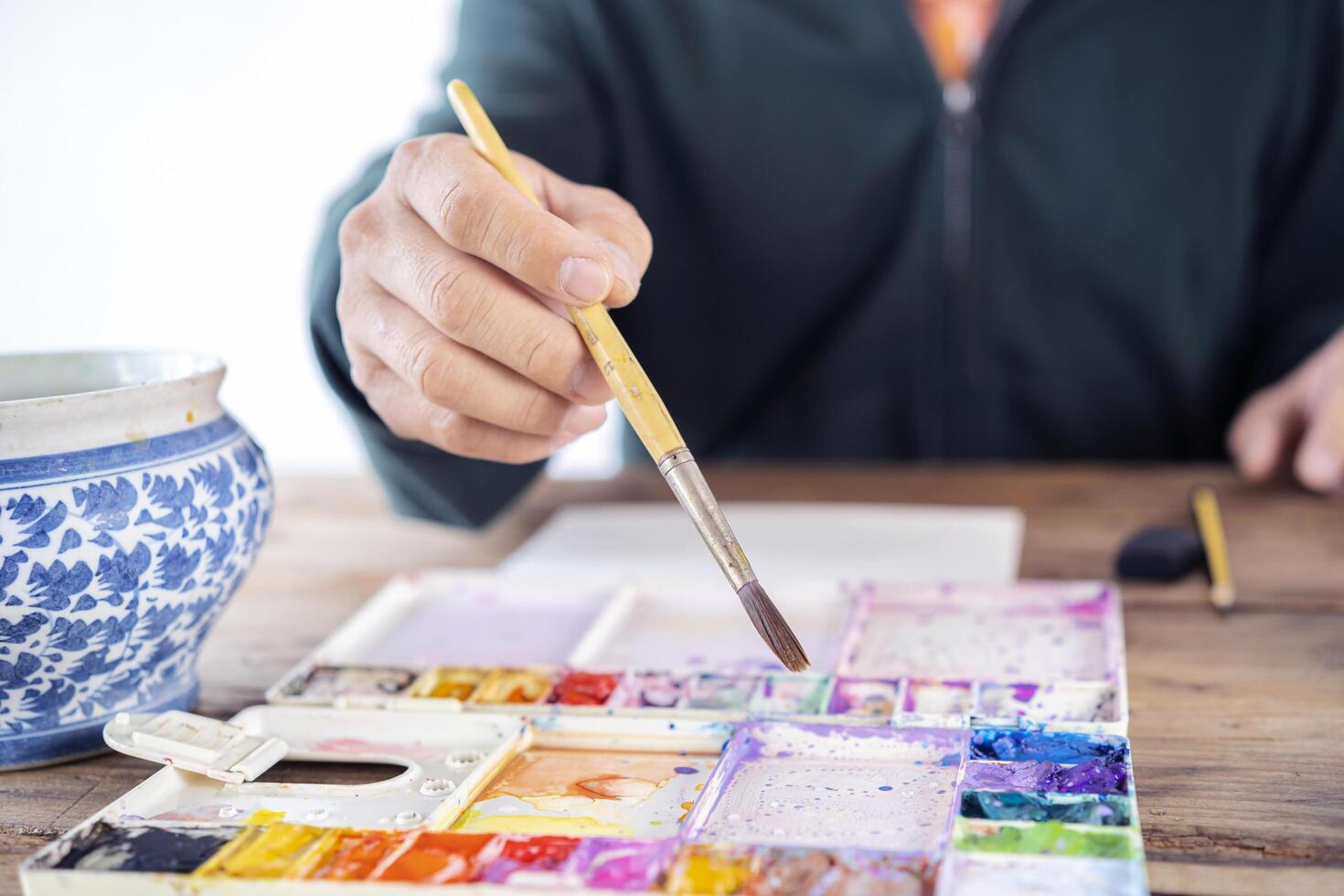 Man artist painting with paint brush and watercolor palette on paper in front of laptop photo