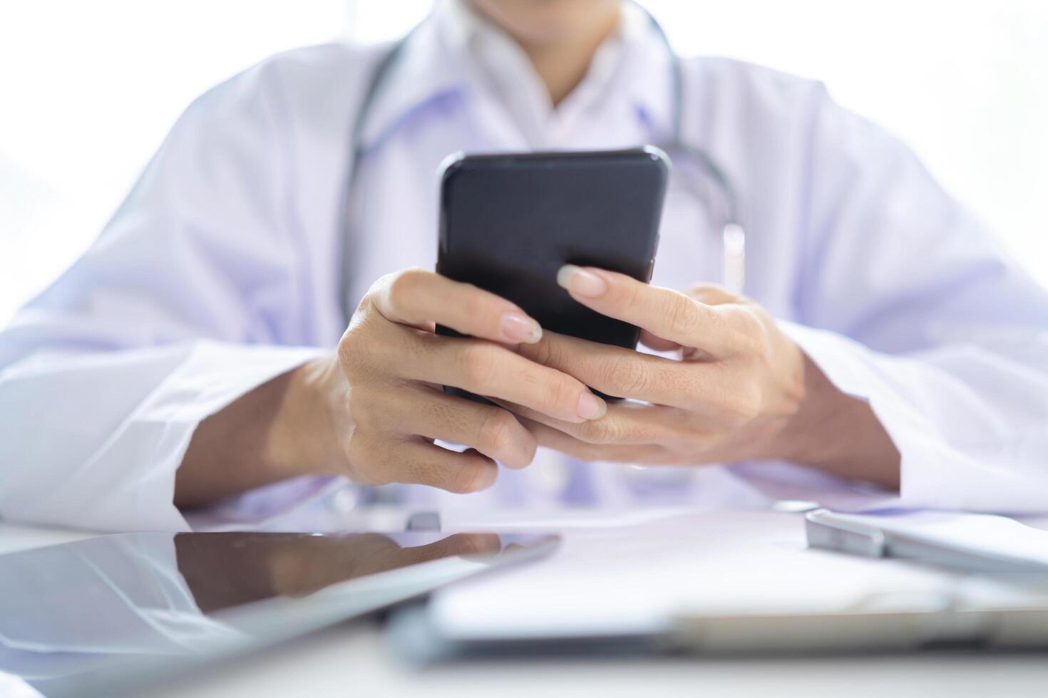 Female doctor hands holding and using mobile smart phone with laptop computer and stethoscope on the table photo
