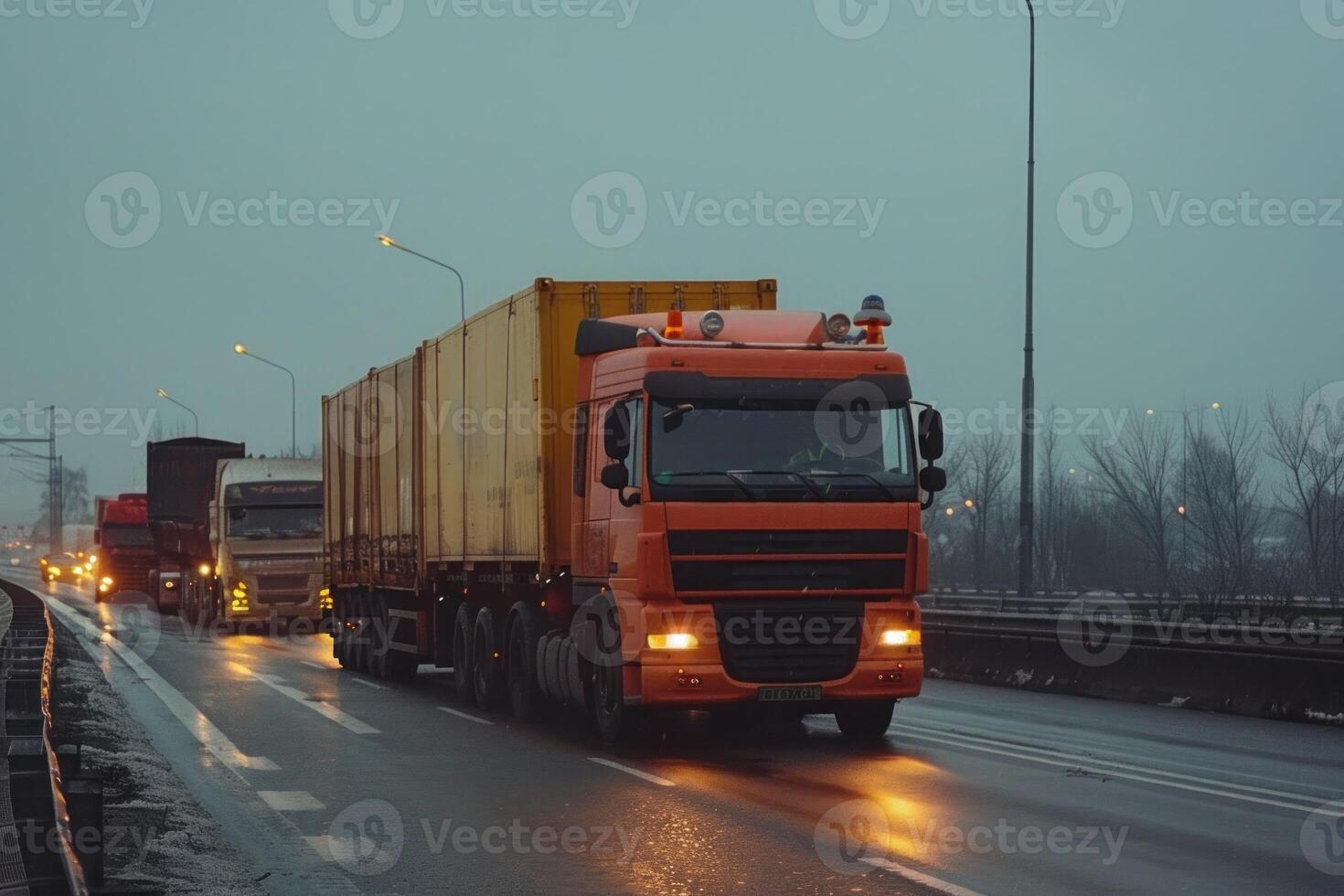 Orange semi truck driving on a highway in rainy weather with other trucks following photo