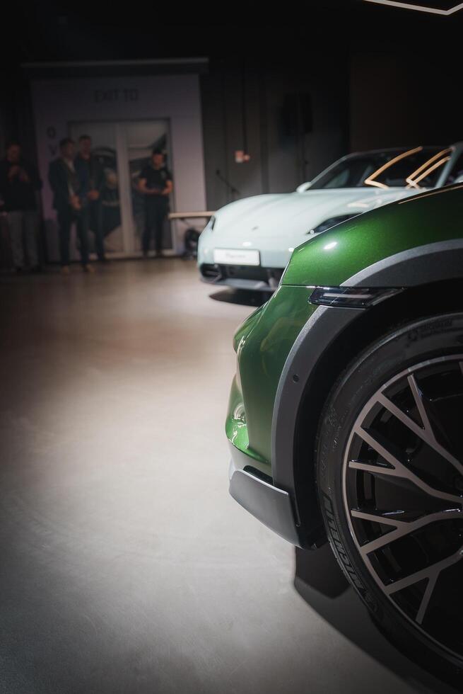 Luxury green car close up showcasing modern wheel design and sleek lines in an upscale indoor setting with blurred white car and people in the background. photo