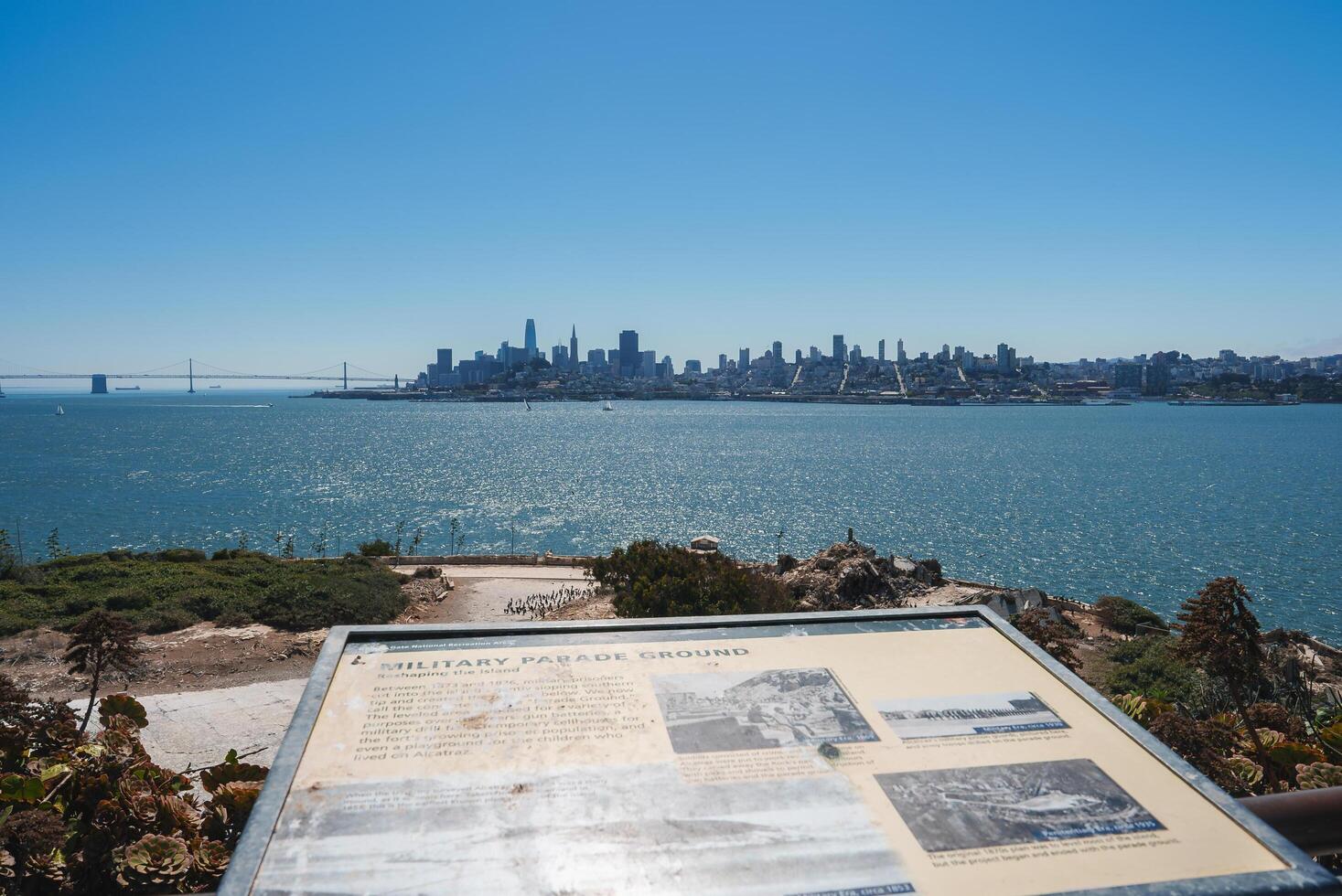 Scenic view of Alcatraz Island with MILITARY PARADE GROUND plaque, looking towards San Francisco skyline. Bay Bridge, boats visible. Sunny day, clear sky, no people. photo