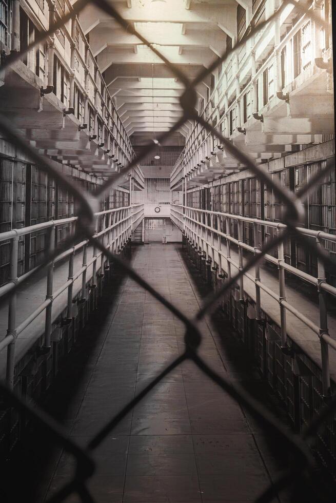 Interior view of Alcatraz prison in San Francisco, USA. Perspective through chain link fence shows cell blocks, barred doors, and stark lighting. Conveys isolation and historical atmosphere. photo