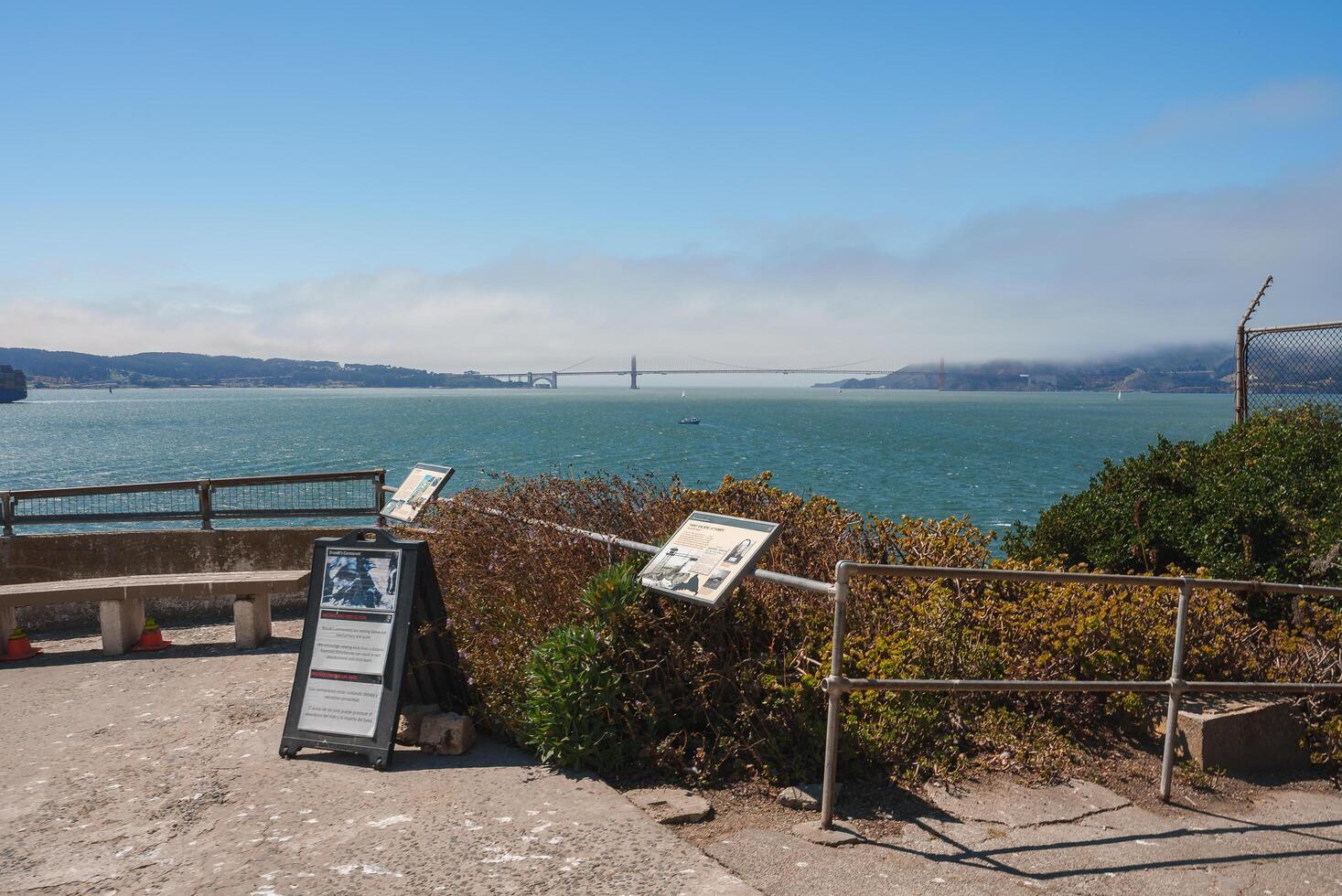 Scenic viewpoint from Alcatraz Island overlooking San Francisco Bay. Metal railing, informational signs, Golden Gate Bridge in misty background, lush vegetation. photo