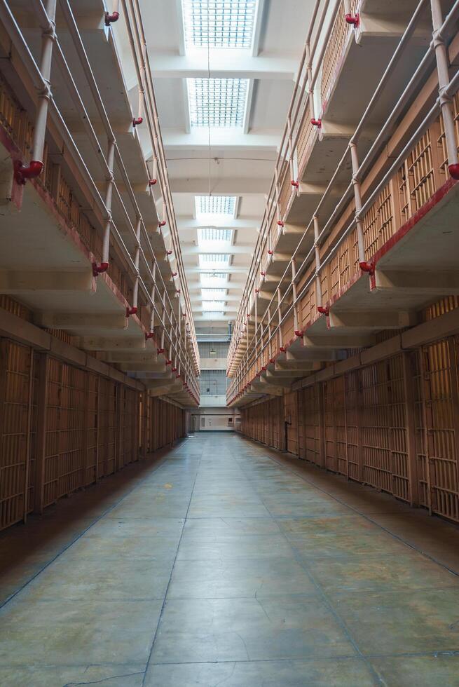 Interior view of Alcatraz prison, San Francisco, USA. Ground level perspective of cell block with barred doors, red pipes, worn green concrete floor, and windows for natural light. photo
