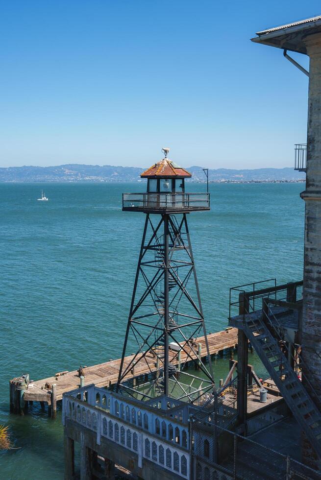 View from Alcatraz prison in San Francisco, USA. A guard tower stands on a pier with a weathered dome roof. A boat floats in the bay with a clear sky. photo