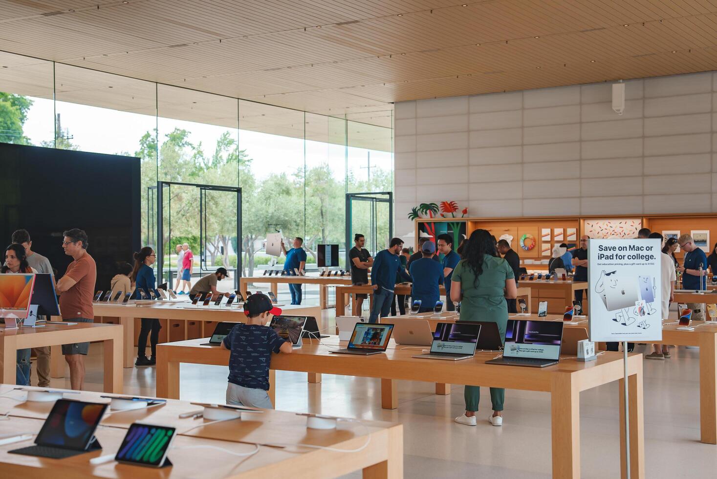 Modern, minimalist Apple Store in Mountain View, California. Wooden tables display MacBooks, iPads, and iPhones. Customers browse while employees assist. Promo sign hints at special deals. photo