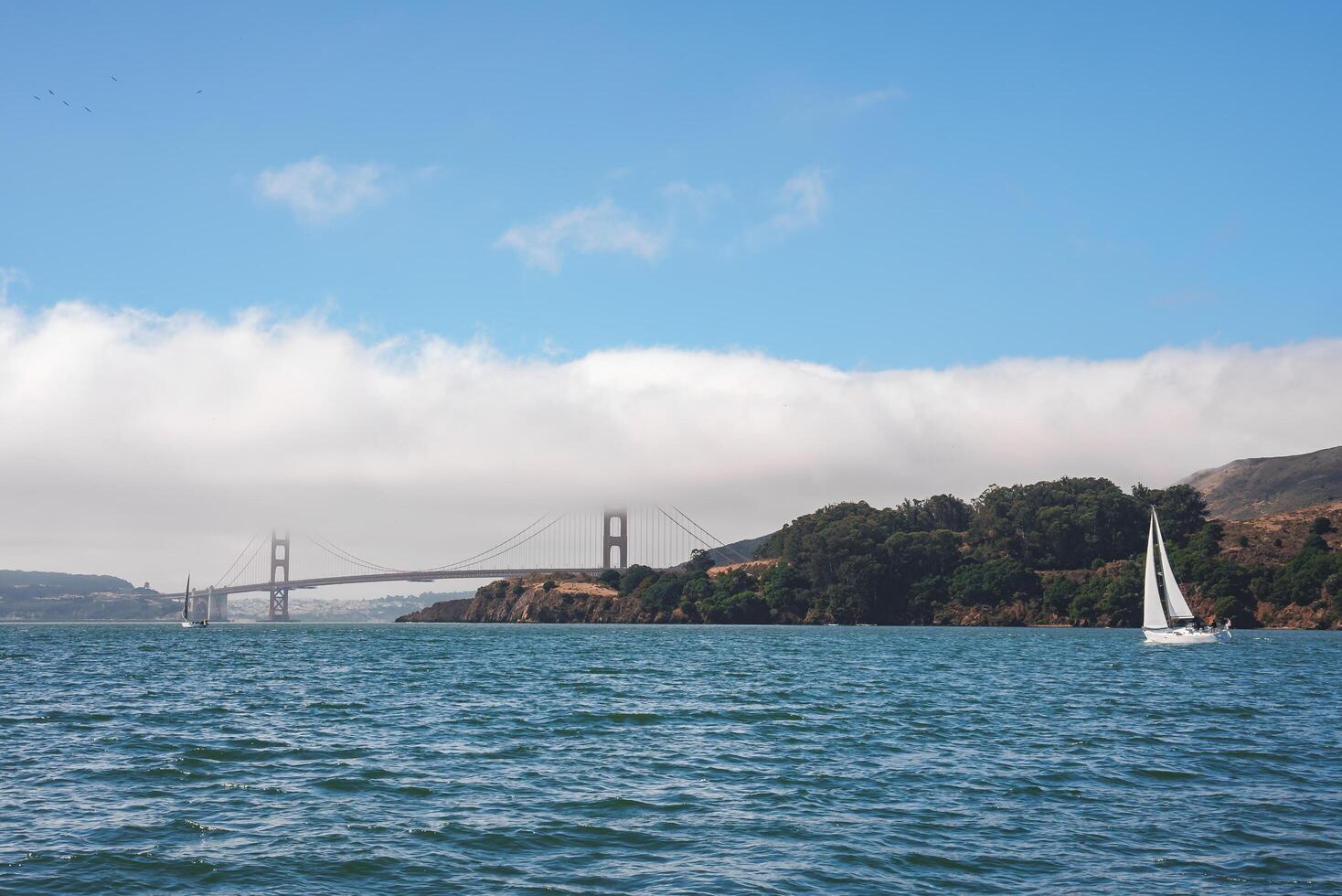 Serene view of Golden Gate Bridge over San Francisco Bay. White sailboat glides on calm water, hills covered in greenery, clear sky with scattered clouds. photo