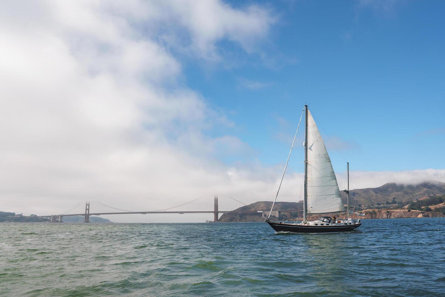 Enjoy a serene maritime view with the iconic Golden Gate Bridge in San Francisco. A yacht glides gracefully with hoisted sails on calm waters, under a clear sky. photo