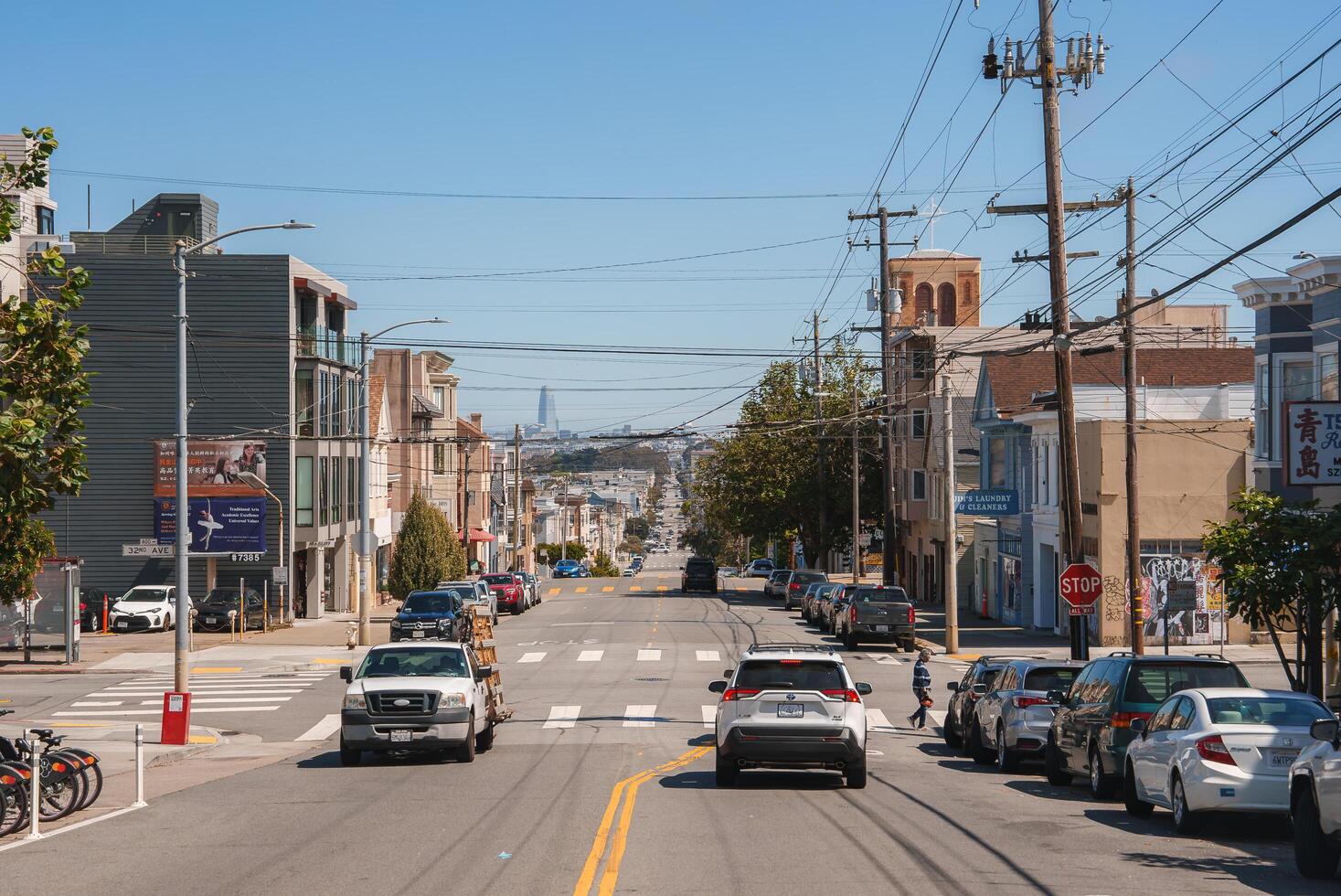 San Francisco street scene featuring iconic bay windows and diverse architecture. Cars drive on sloping road with clear blue sky and utility lines in view. photo