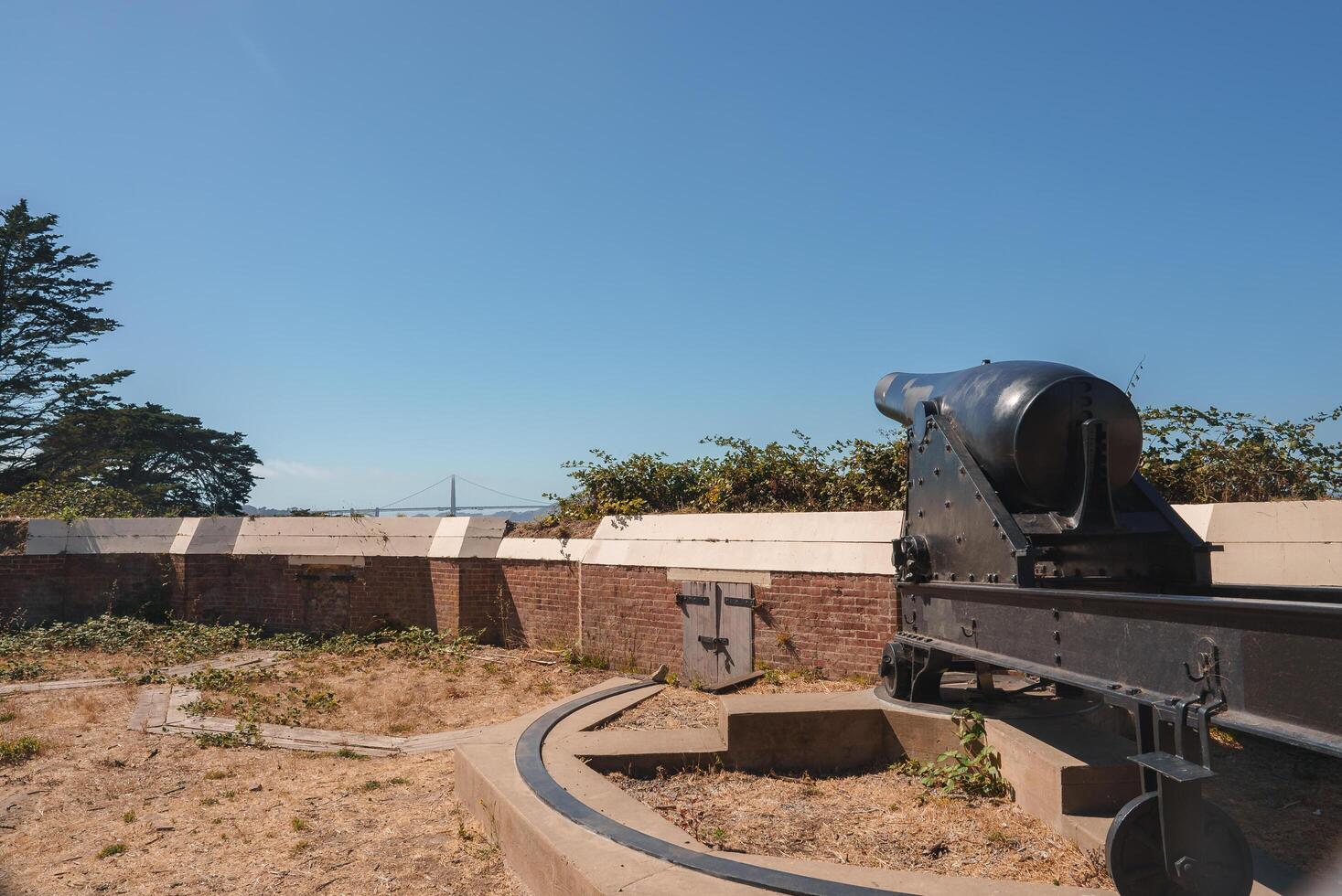 An old cannon on a metal carriage sits on sandy ground, with sparse vegetation, a brick wall, and water view. Possibly at a historic site in San Francisco. photo