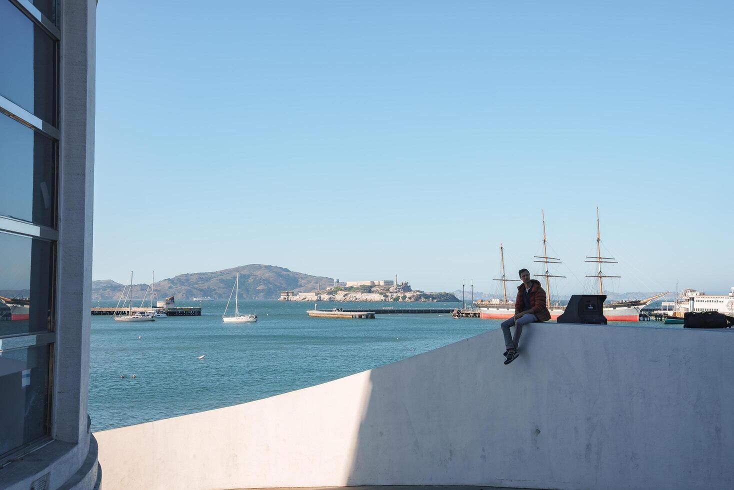 Serene waterfront view in San Francisco person with laptop on ledge, boats in bay, calm water reflecting blue sky, city backdrop, tranquil atmosphere. photo