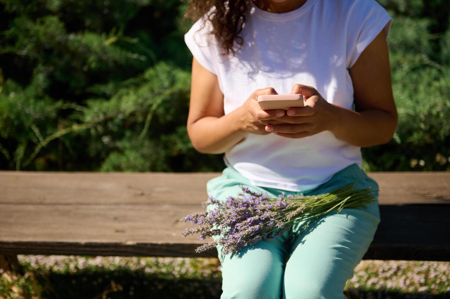 Woman using smartphone while sitting on park bench with lavender flowers in summer day photo
