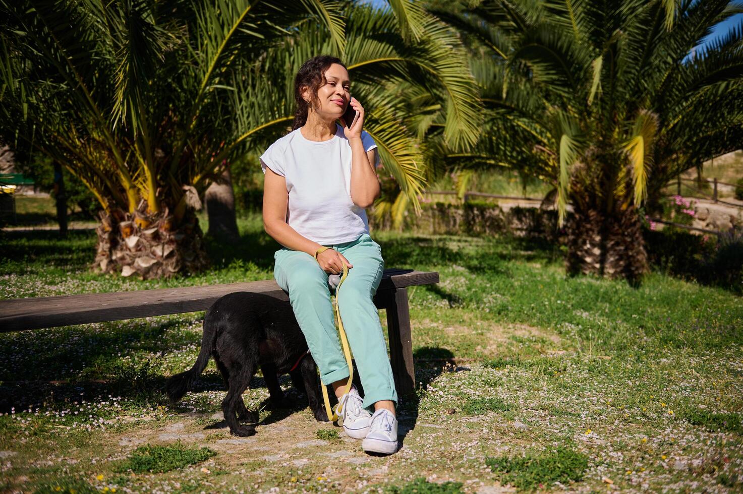 Woman enjoying phone conversation while sitting on park bench with her dog on a sunny day photo
