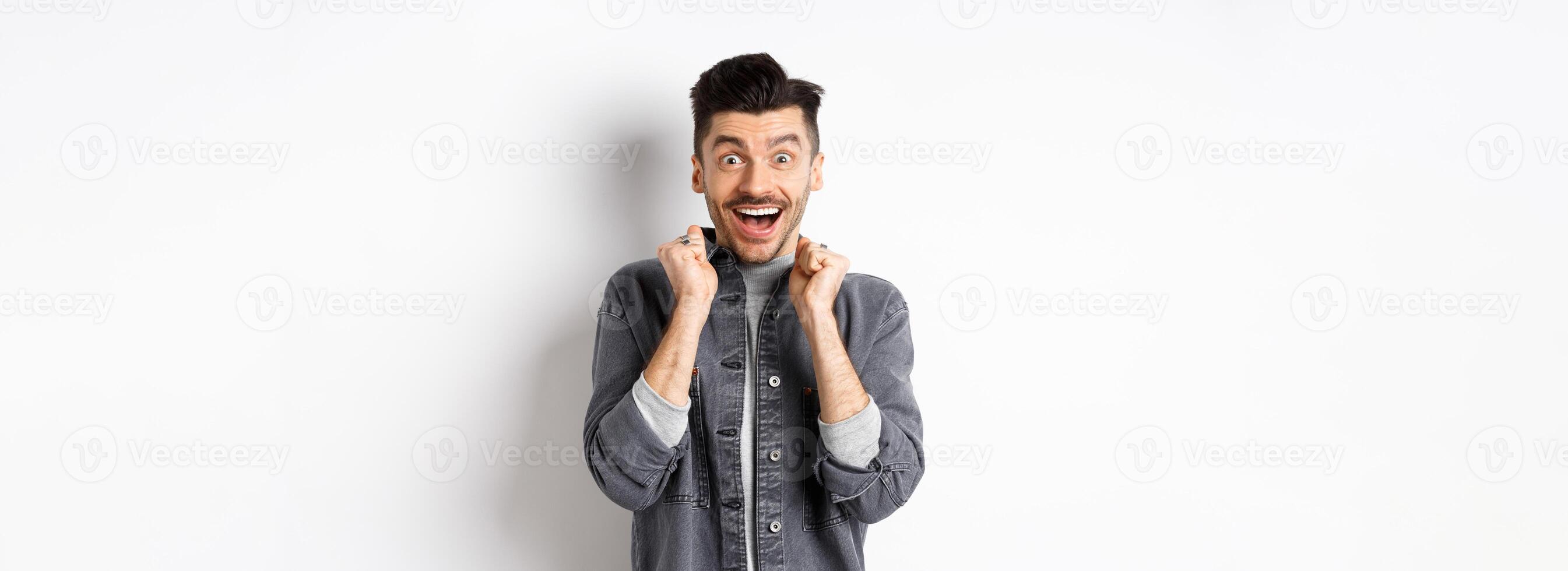 Excited young man jumping from happiness, smiling amazed and holding hands pressed to chest, standing on white background photo