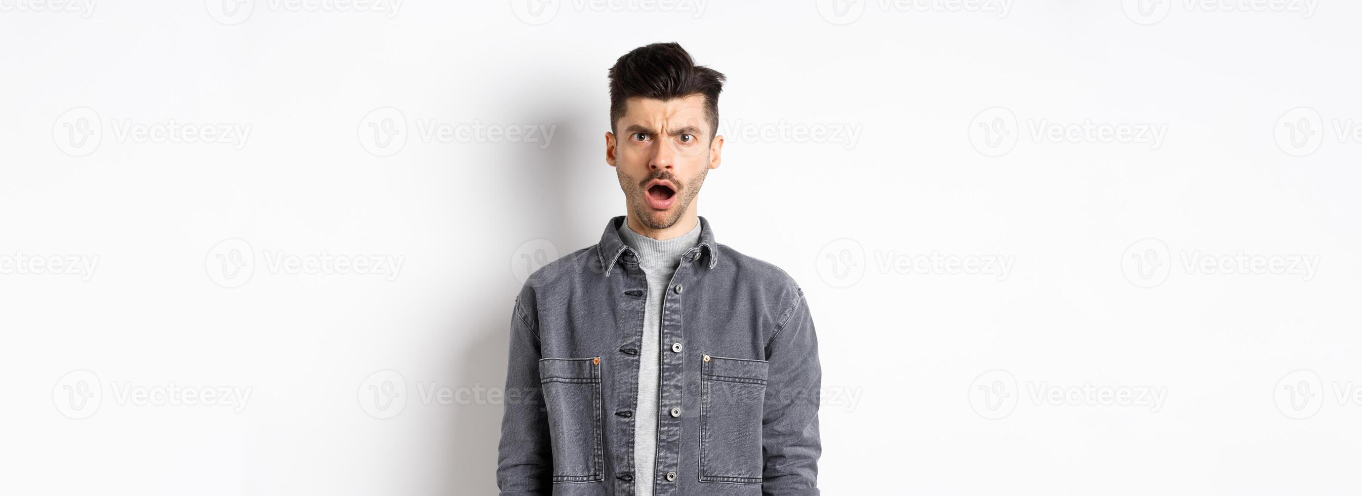 Shocked and offended man frowning, looking at camera with opened mouth, feeling hurt by cruel words, standing disappointed on white background photo