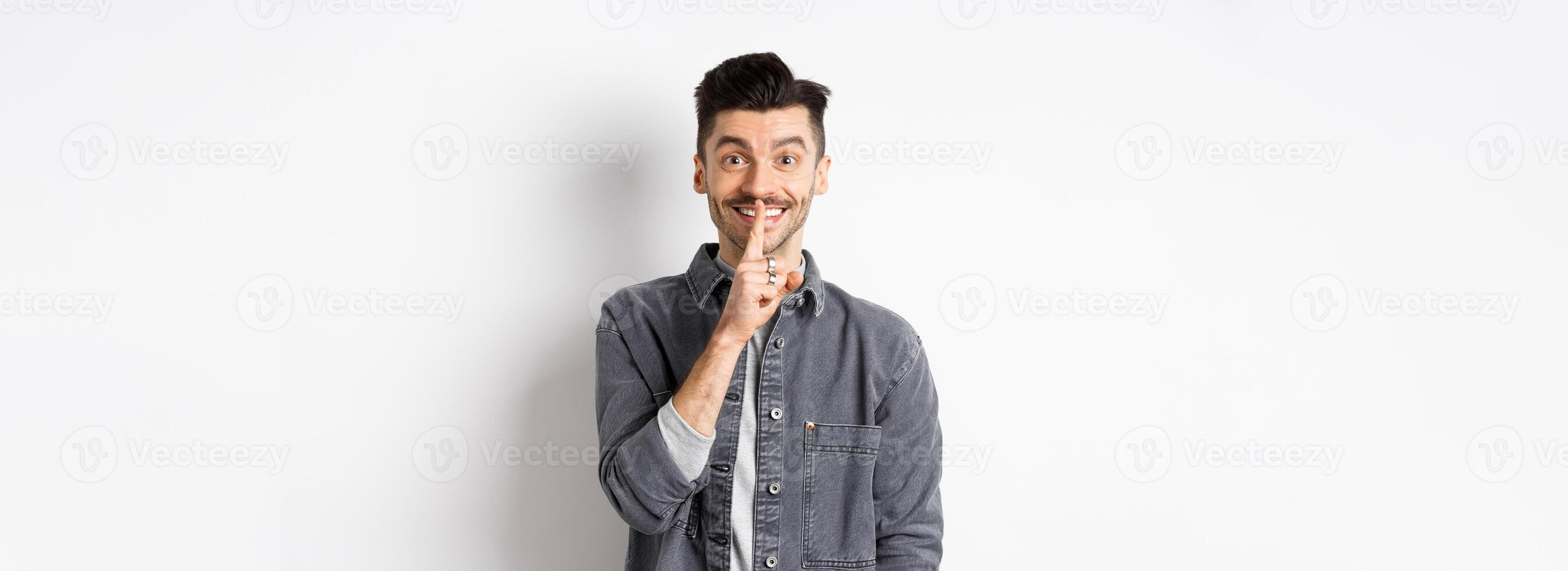 Happy guy shushing at camera, smiling with finger on lips, asking to keep quiet, telling big secret or making surprise, standing on white background photo