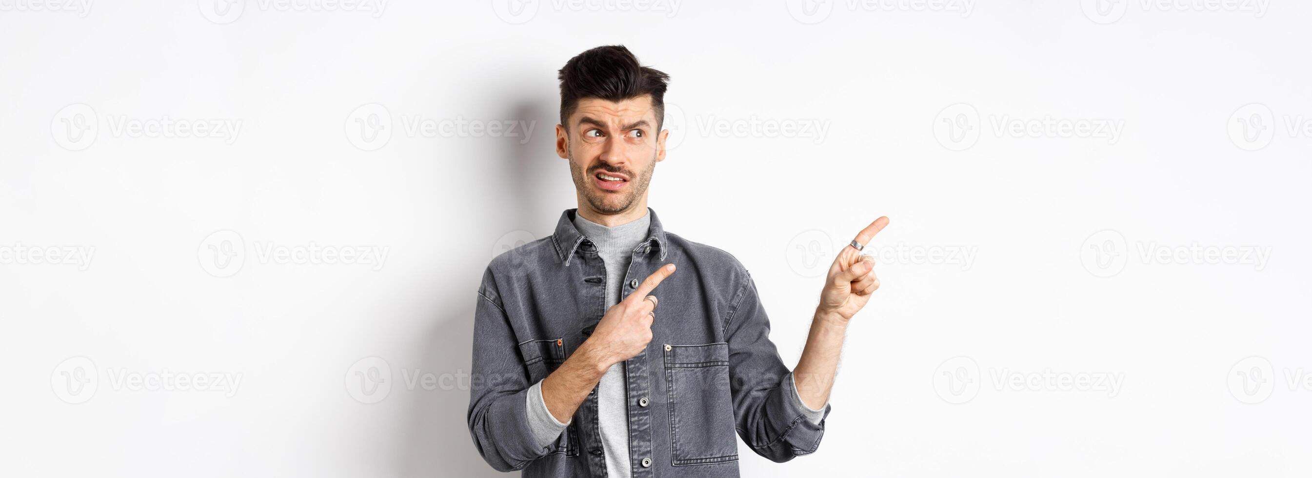Skeptical and doubtful young man pointing fingers left at logo, look with dislike and confusion, standing on white background photo