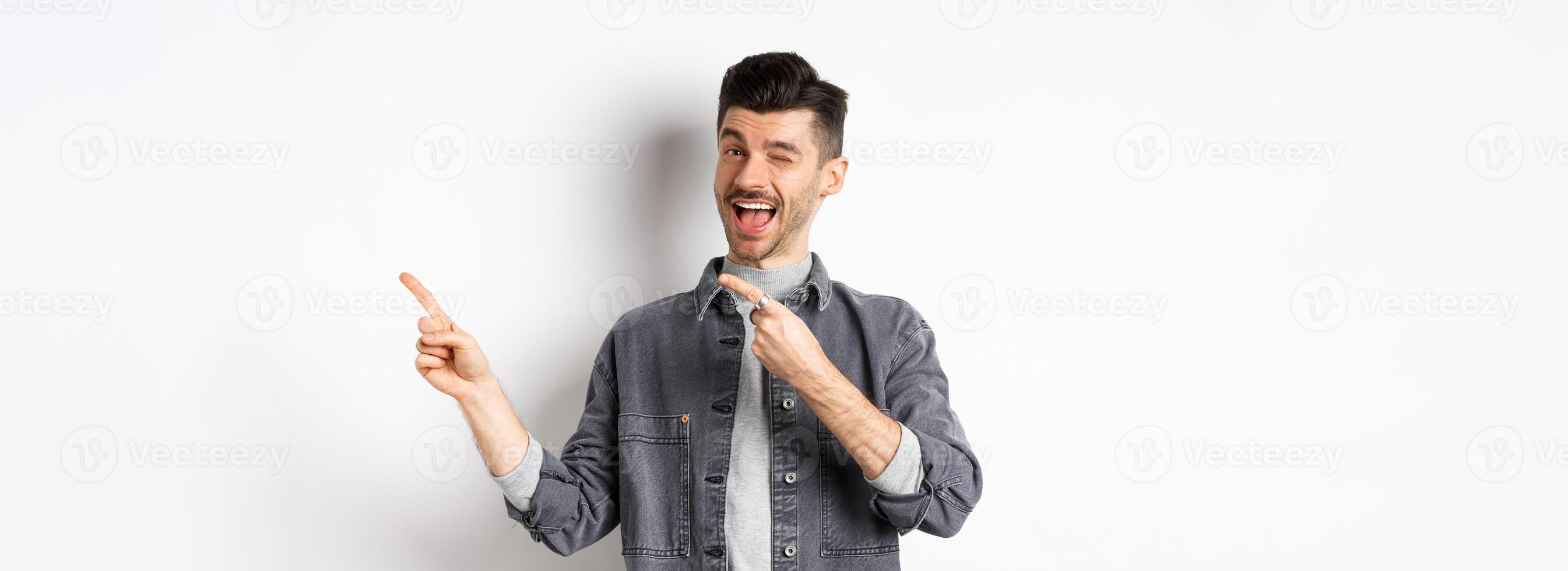 Handsome smiling guy making announcement, winking and pointing at upper right corner, showing logo, inviting to check out advertisement, standing on white background photo