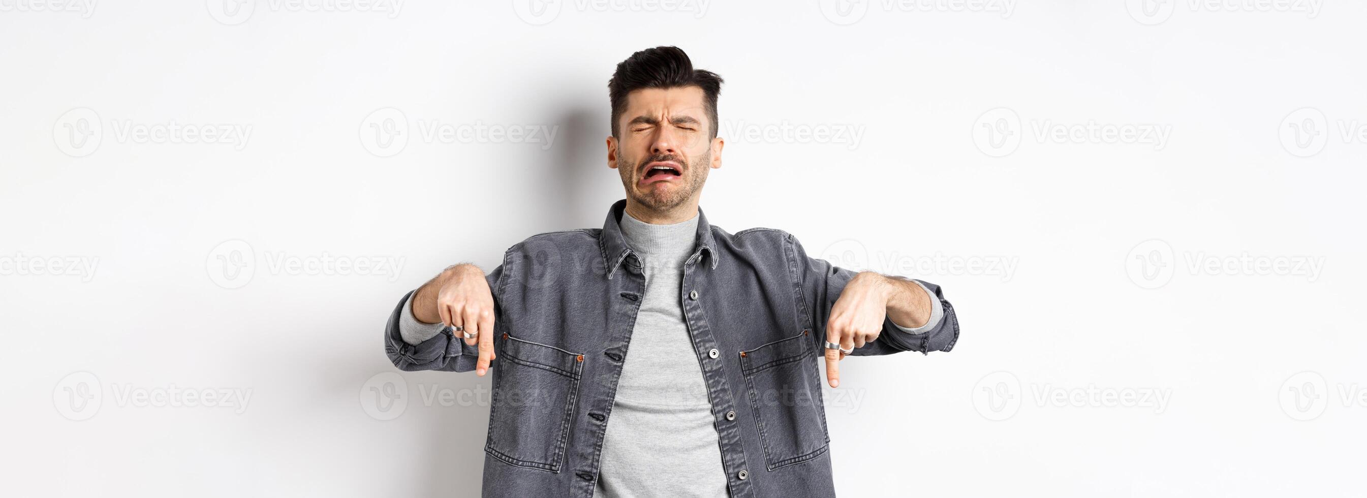 Sad crying man pointing fingers down, sobbing miserable, showing upsetting news, standing in denim jacket on white background photo
