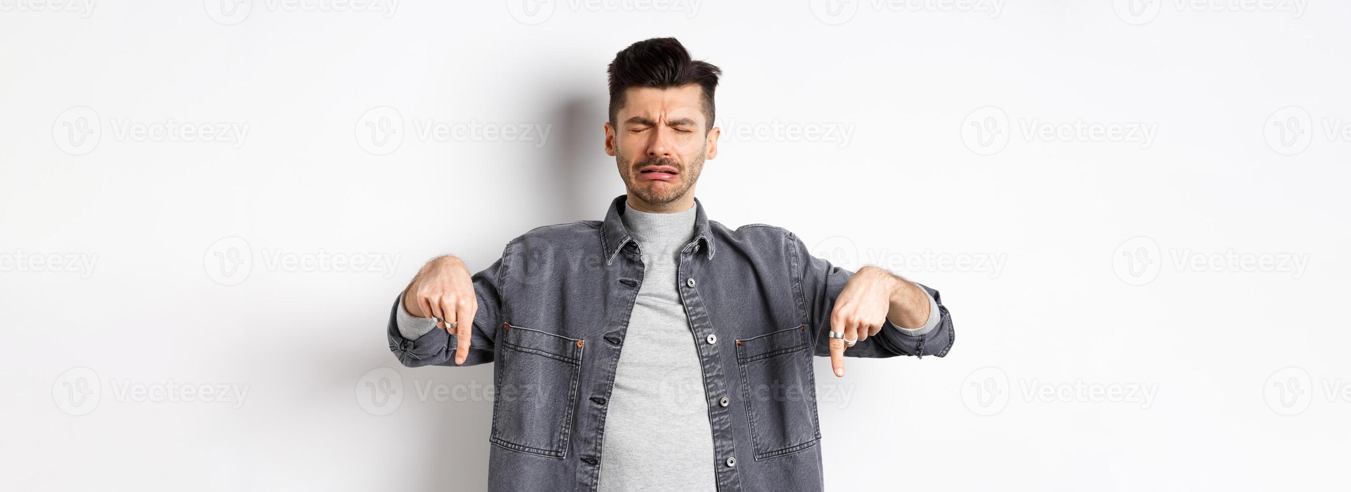 Sad crying man whining, pointing fingers down disappointed, showing bad news, standing upset on white background photo