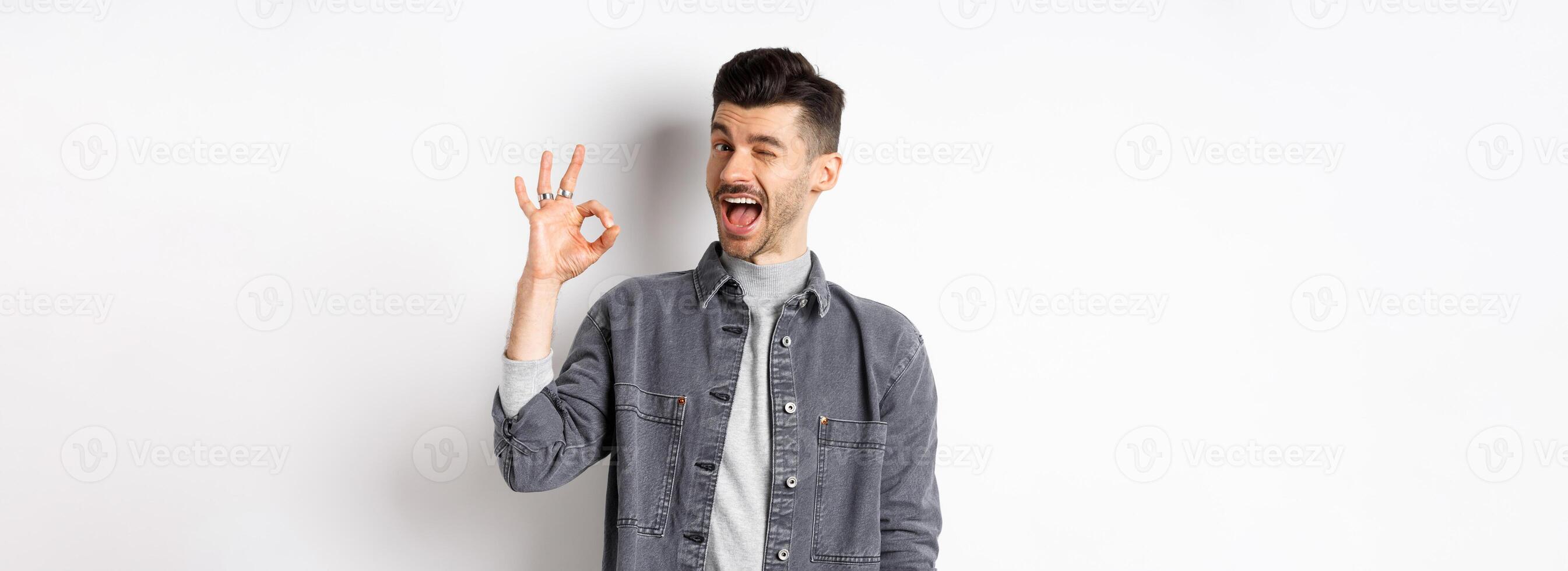 Excited handsome guy with moustache, winking and showing okay sign, smiling pleased, assure all good, praise nice work, making good job gesture, standing against white background photo