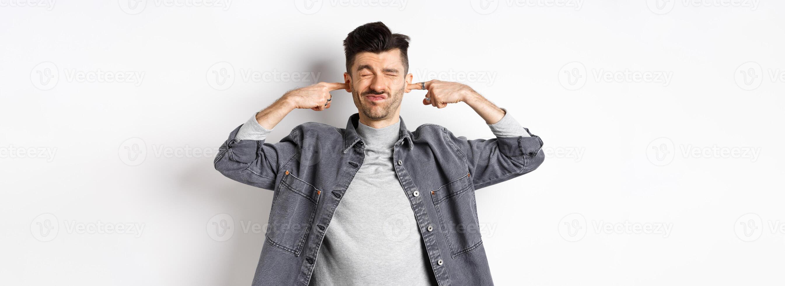 Annoyed young man plug ears with fingers and grimacing from loud noise, disturbed by noisy neighbours, standing at loud place on white background photo
