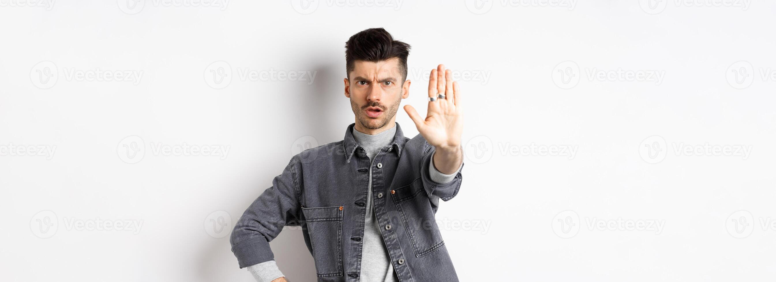 Serious man tell to stop, prohibit something and frowning, say no, standing displeased with stretch out hand against white background photo