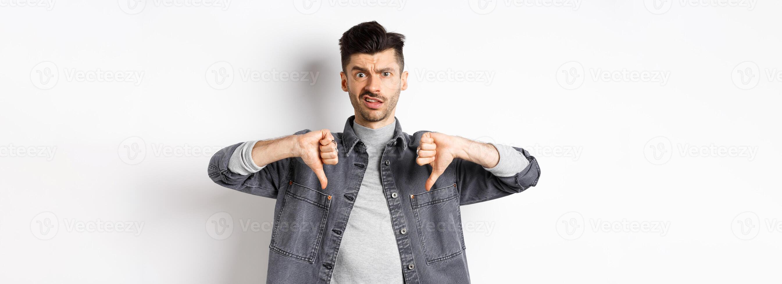 Disappointed guy showing thumbs down and frowning, looking skeptical at something bad, dislike and disapprove awful thing, standing on white background photo