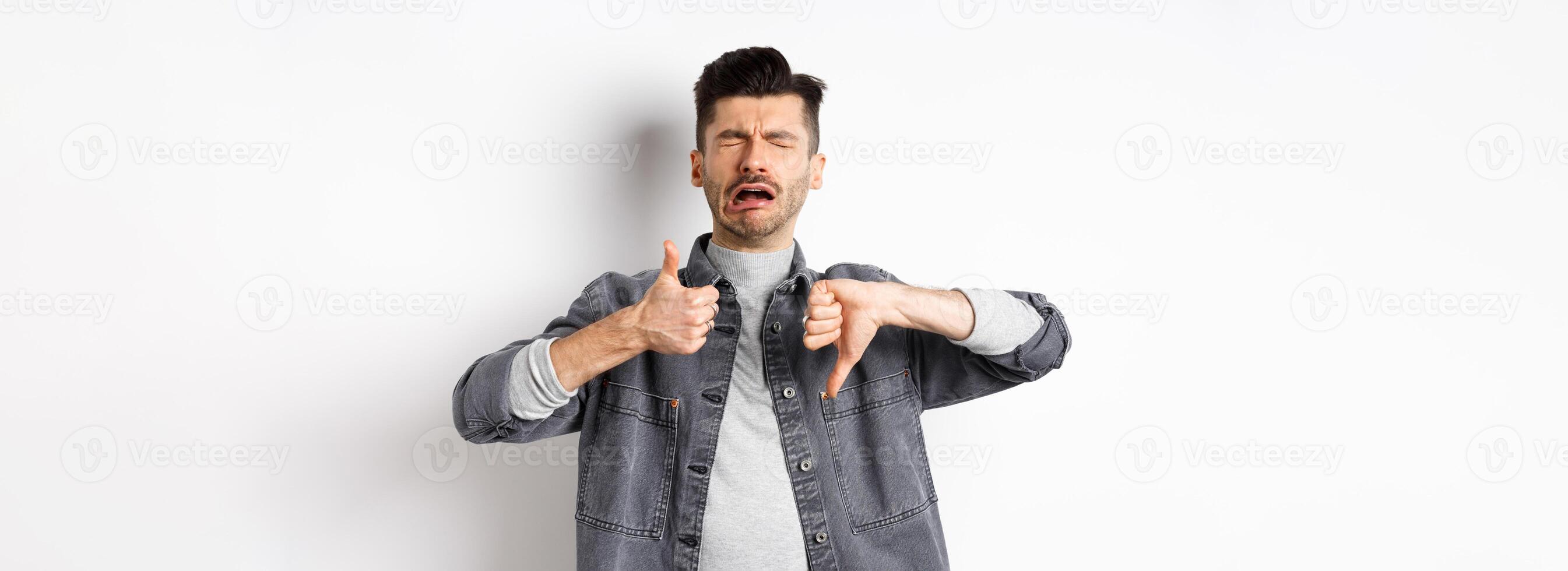 Sad man crying and showing thumbs up down, whining and complaining, standing disappointed against white background photo