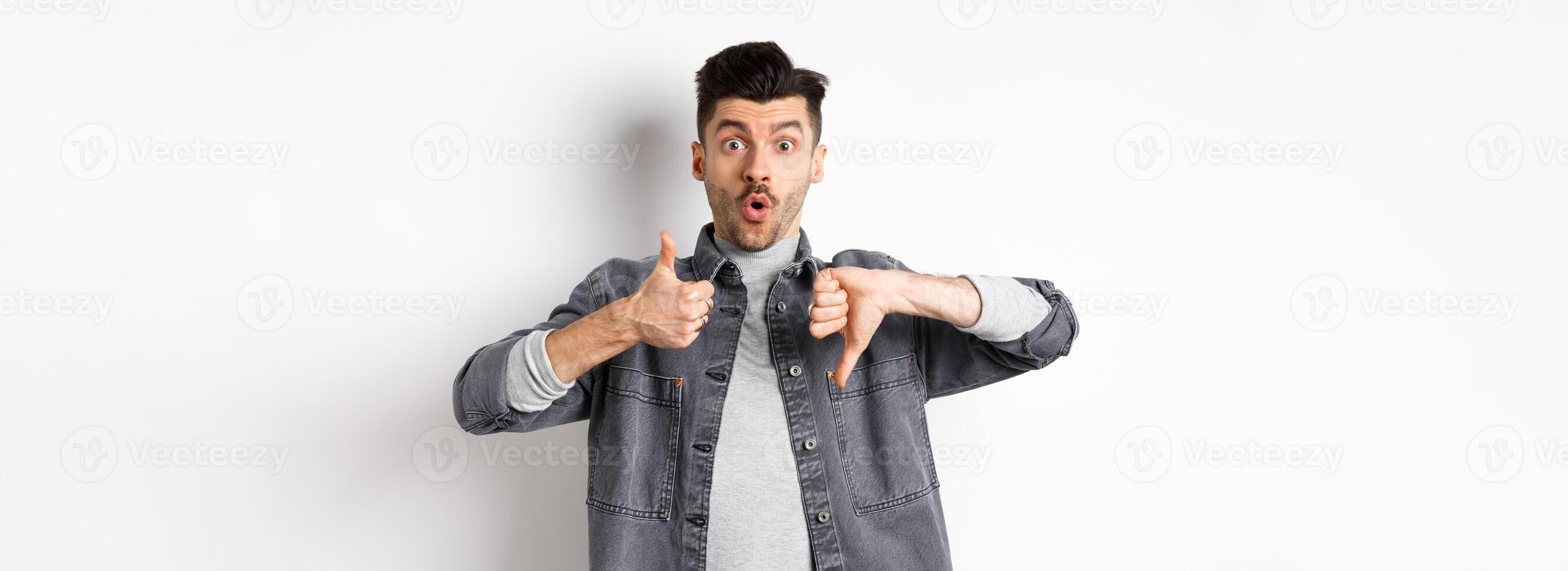 Excited guy show thumbs up and down judging and making decision, rate something interesting, standing on white background photo
