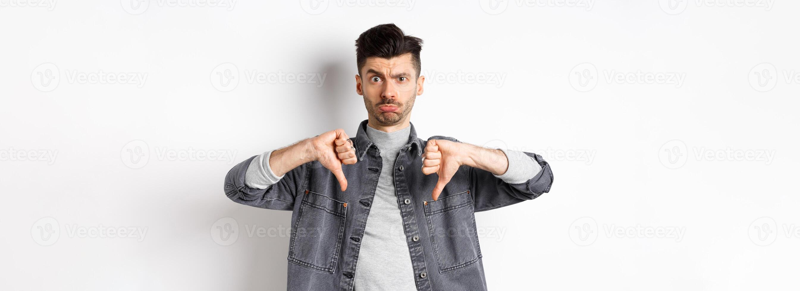 Disappointed funny man frowning and looking upset, showing thumbs down displeased, standing on white background photo