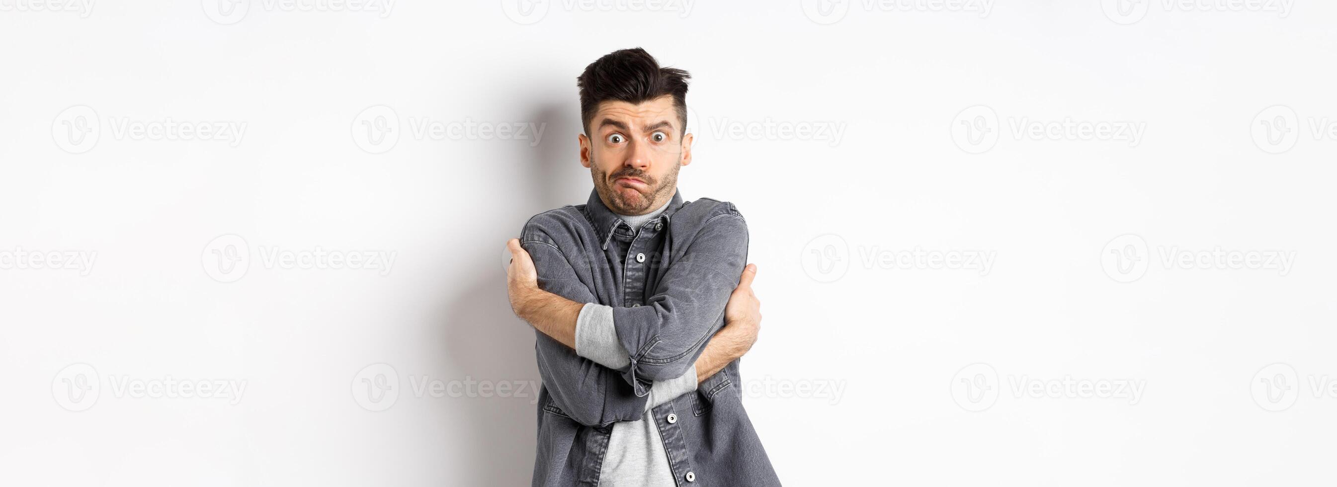 Funny young man hugging himself and grimacing, feeling embarrassed and exposed, covering body from people glances, white background photo