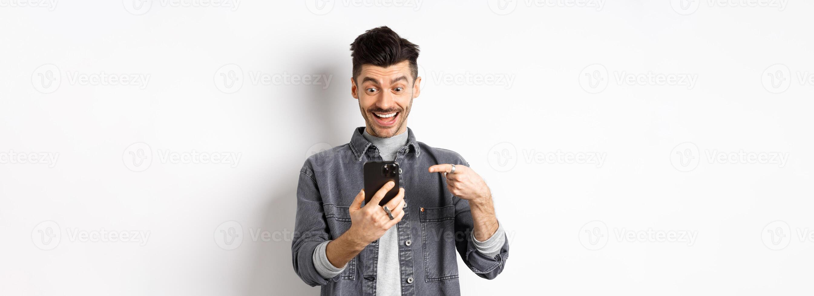 Excited handsome man showing cool news on phone, pointing at smartphone and smiling happy, standing against white background photo
