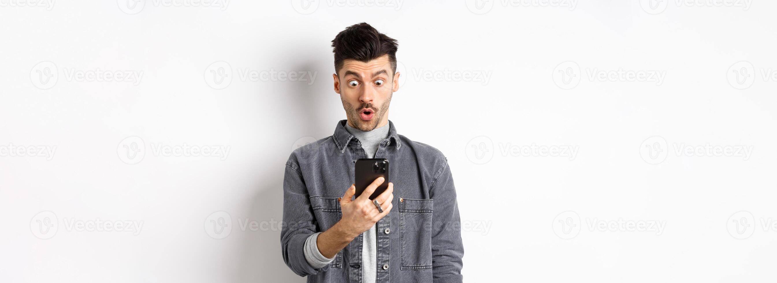 Man look surprised at smartphone screen, receive exciting news on phone, standing against white background photo