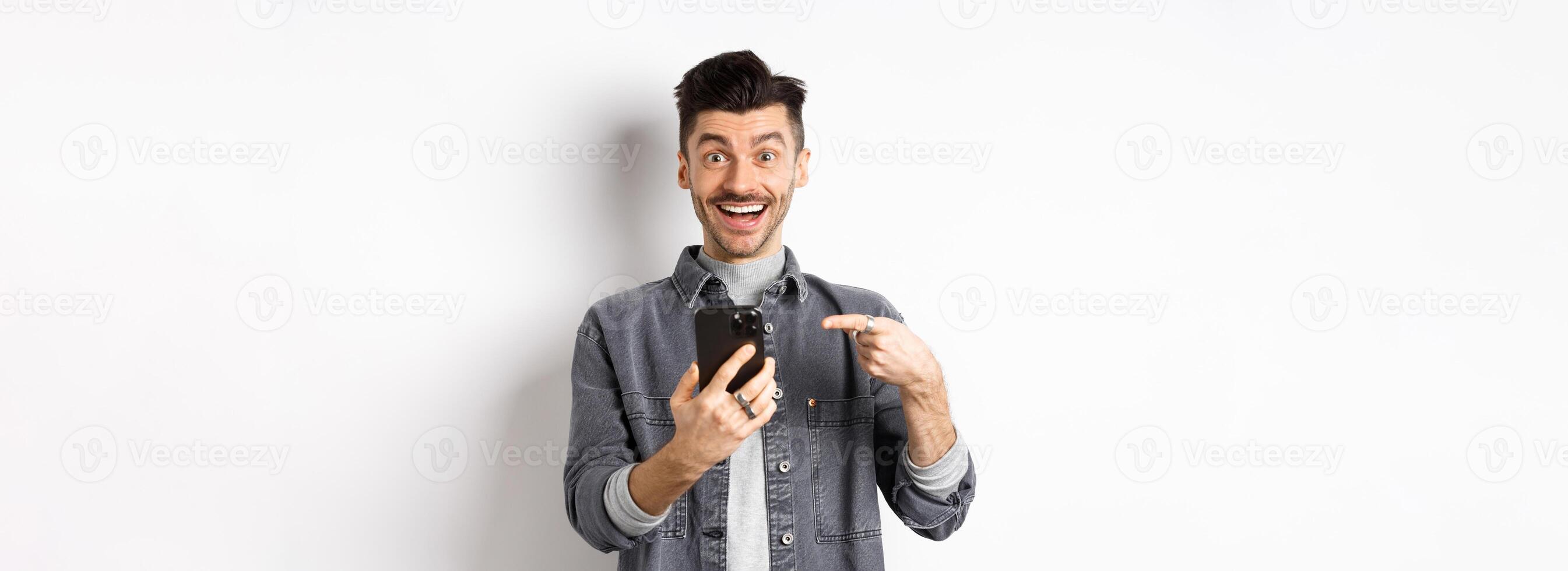 Excited young man receive good news on phone, pointing at smartphone and smiling happy, standing on white background photo