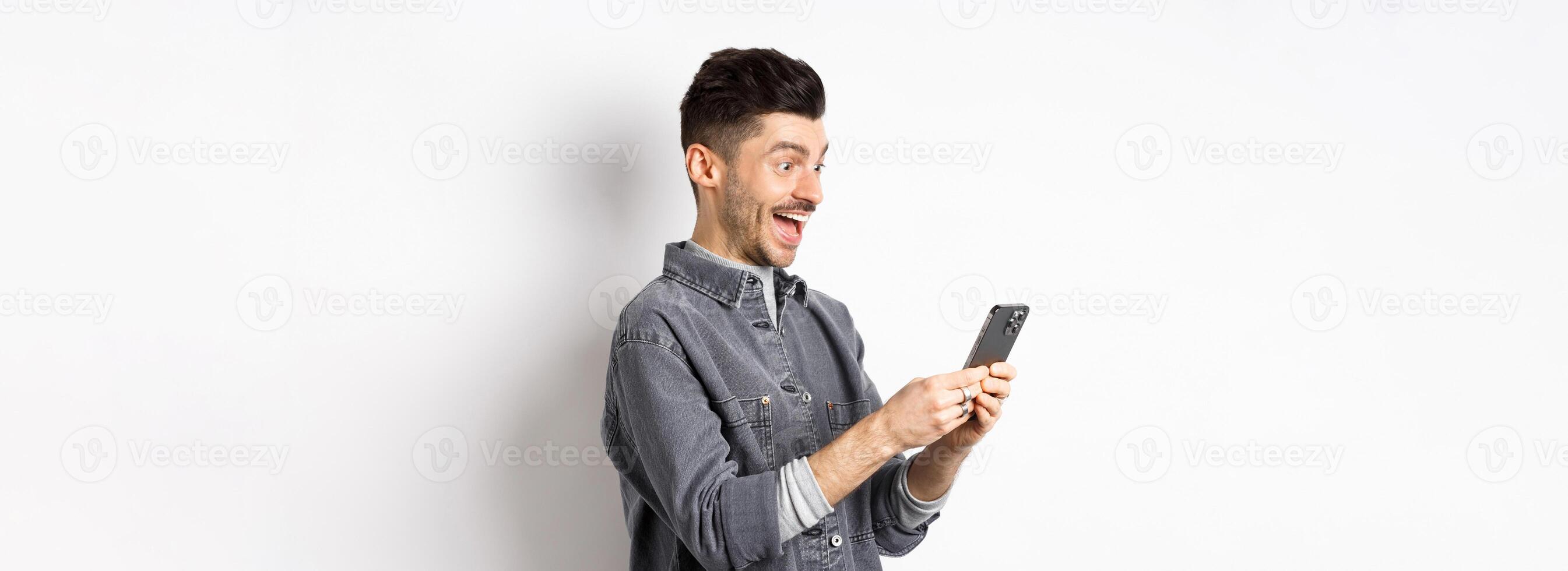Profile portrait of excited handsome man reading message on phone, looking happy at smartphone screen, standing on white background photo