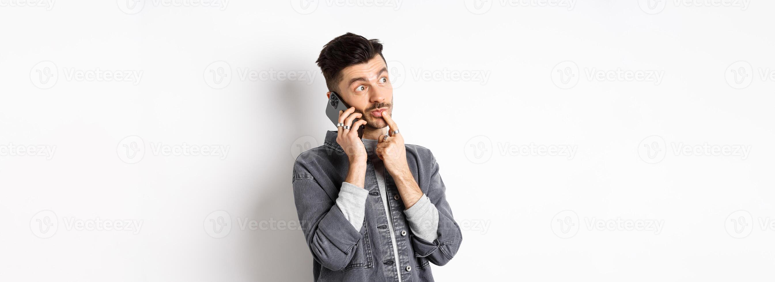 Image of pensive handsome man talking on phone and thinking, making choice while making takeout order, standing against white background photo