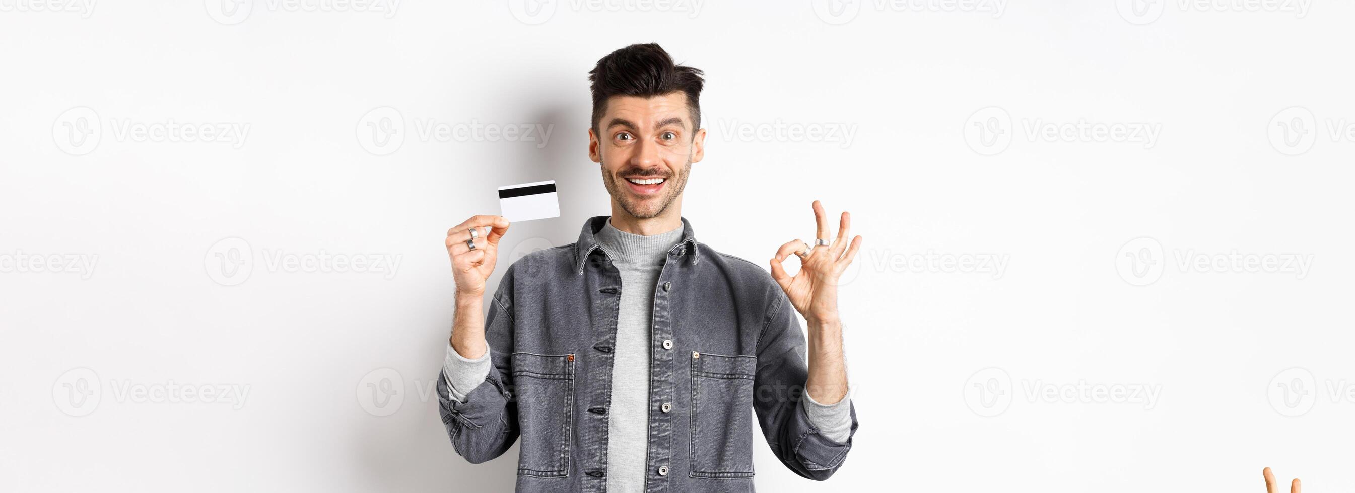Very good. Smiling guy with plastic credit card showing okay sign, smiling satisfied, recommend bank, standing on white background photo