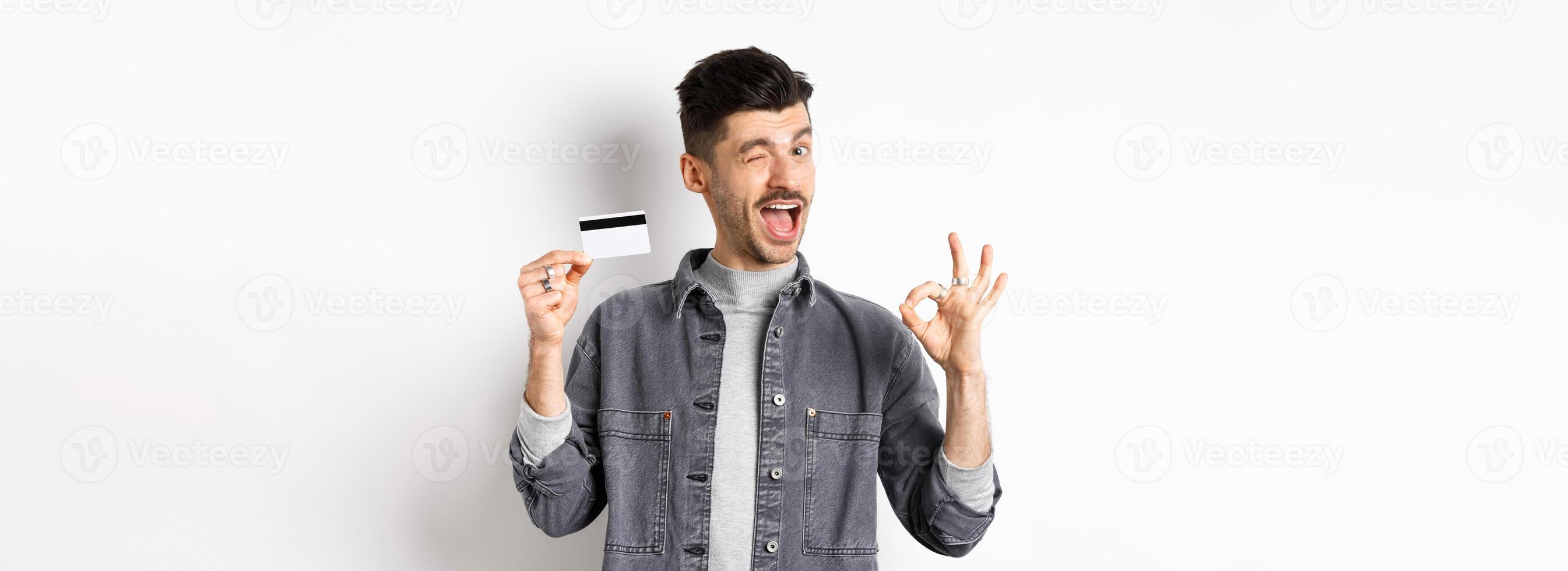 Very good. Smiling guy with plastic credit card showing okay sign, smiling and winking satisfied, recommend bank, standing on white background photo