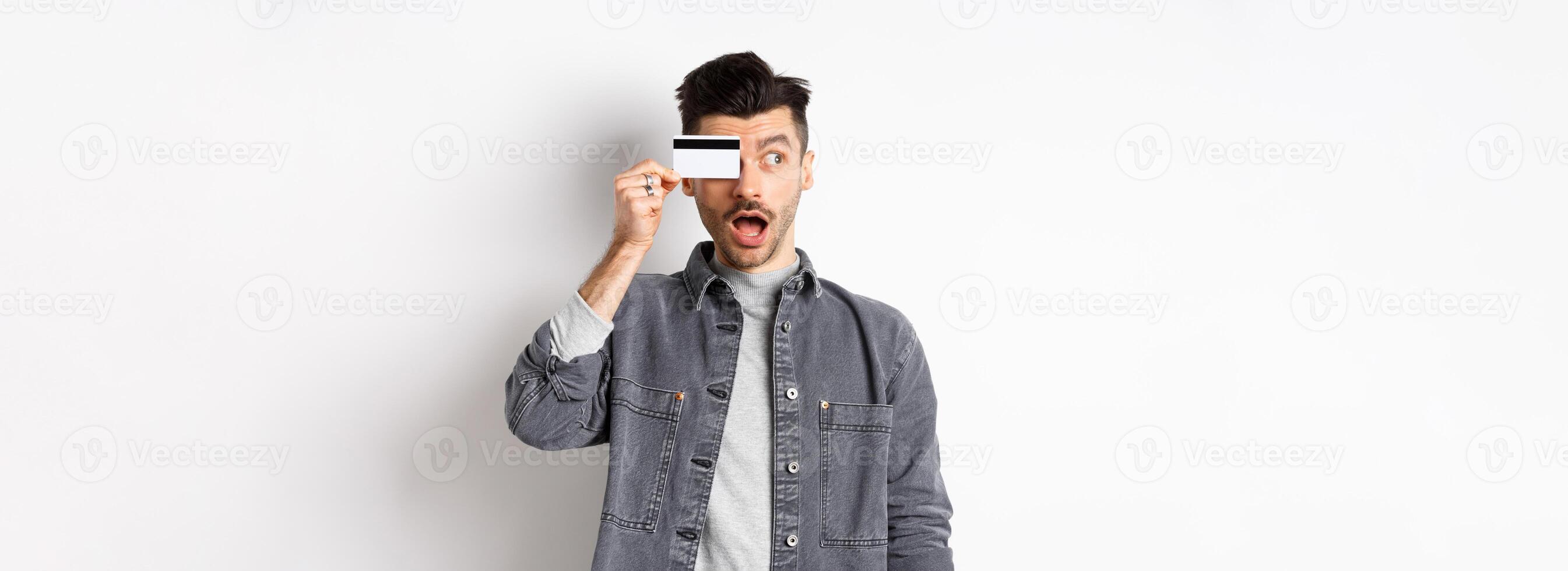 Excited funny guy look aside at logo and show plastic credit card, standing amazed on white background photo