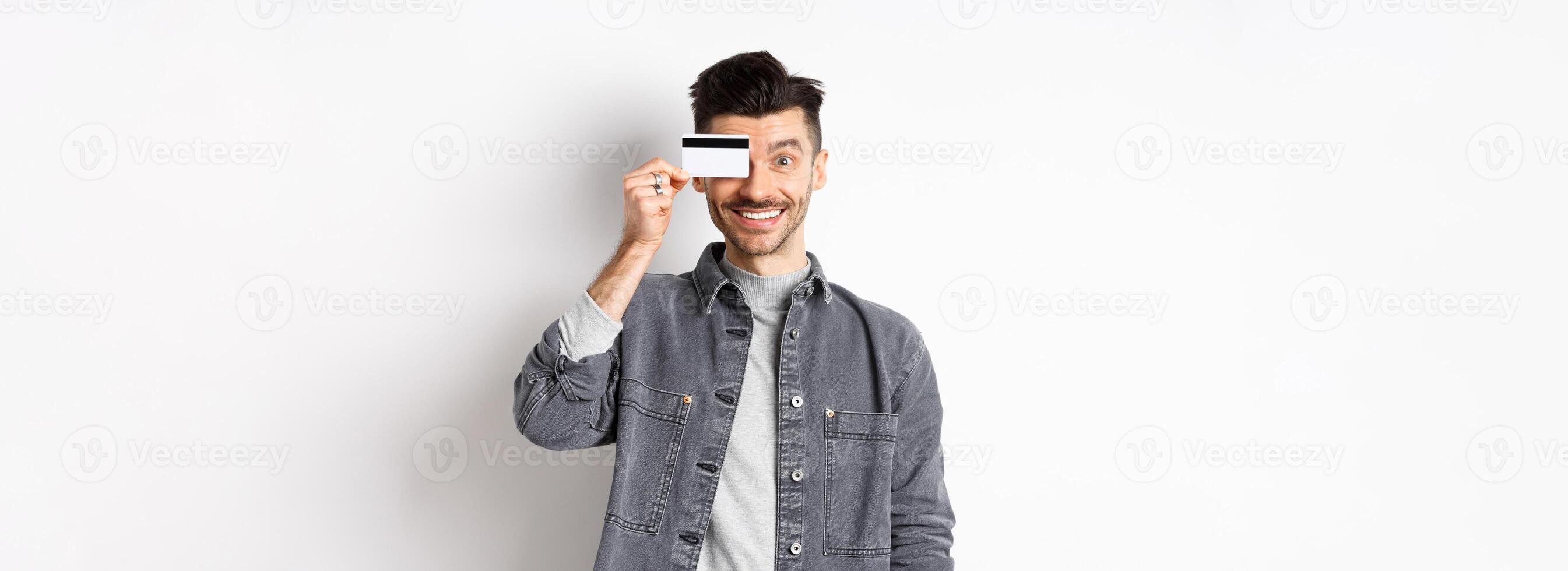 Happy handsome man with moustache show plastic credit card on eye, smiling excited at camera, standing on white background photo