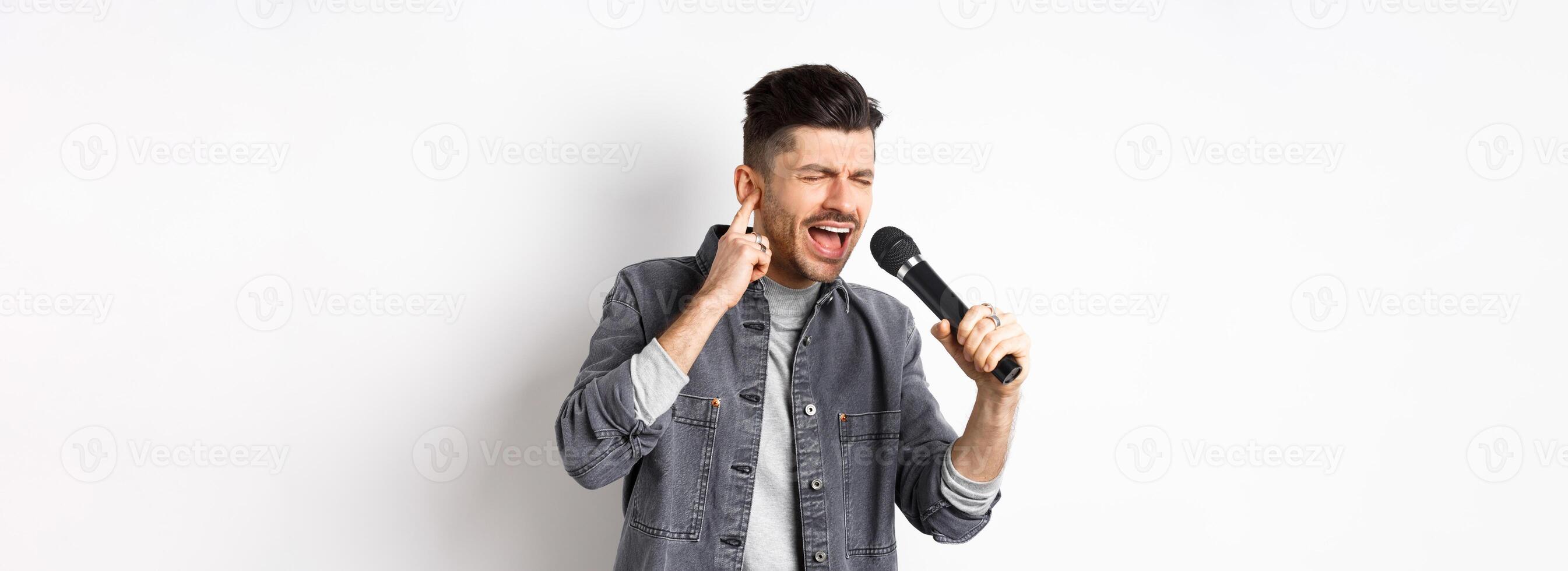 Handsome singer trying to reach high note, singing in microphone and shut one ear, standing on white background photo