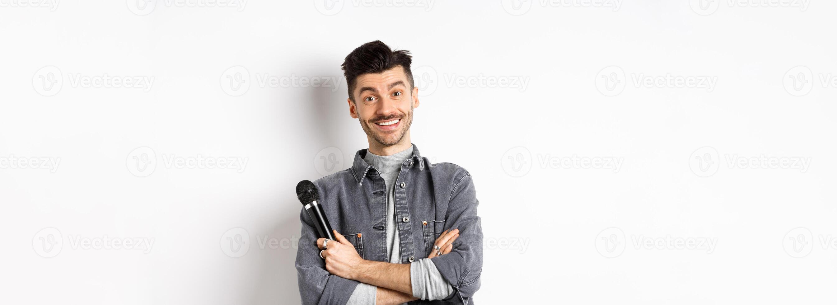 Handsome caucasian guy with moustache cross arms on chest, holding mic and smiling at camera, perform on stage with microphone, standing against white background photo